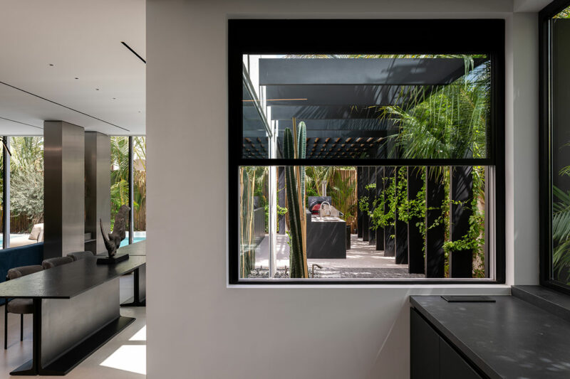 Modern interior with a view through a large window to an outdoor patio featuring black pergolas, lush green plants, and natural sunlight.