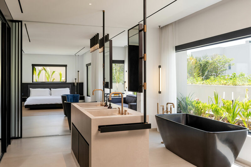 Modern open-concept bedroom and bathroom with a black freestanding tub, double sink, large windows, and views of a garden.