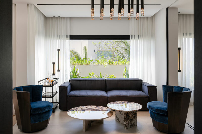 Modern living room with a dark blue sofa, two matching velvet chairs, marble coffee tables, pendant lights, sheer curtains, and a large window overlooking greenery.