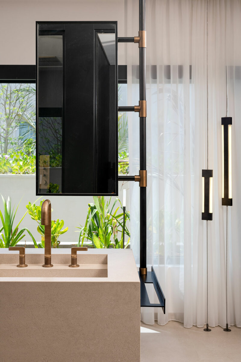 Modern bathroom with a beige stone sink, brass fixtures, a large mirror, floor-to-ceiling window, white curtains, and vertical pendant lights. Green plants visible outside.