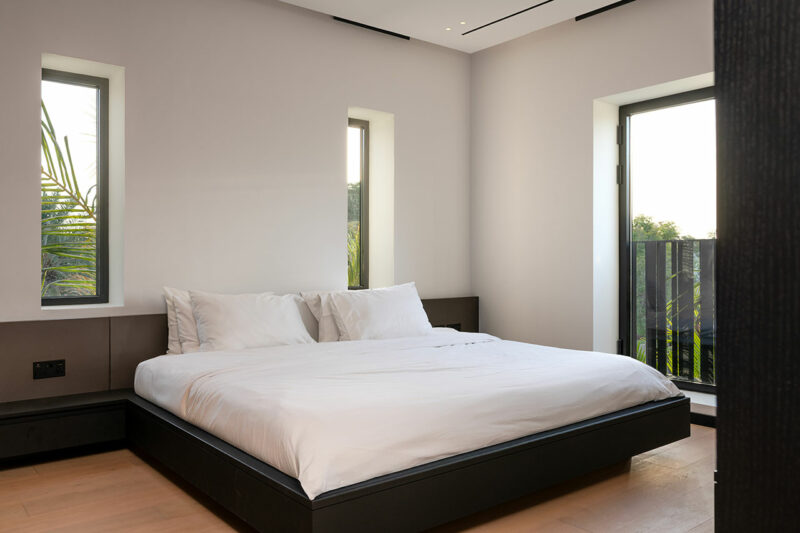 A modern, minimal bedroom with a low platform bed, white bedding, light wood floor, and large windows letting in natural light.