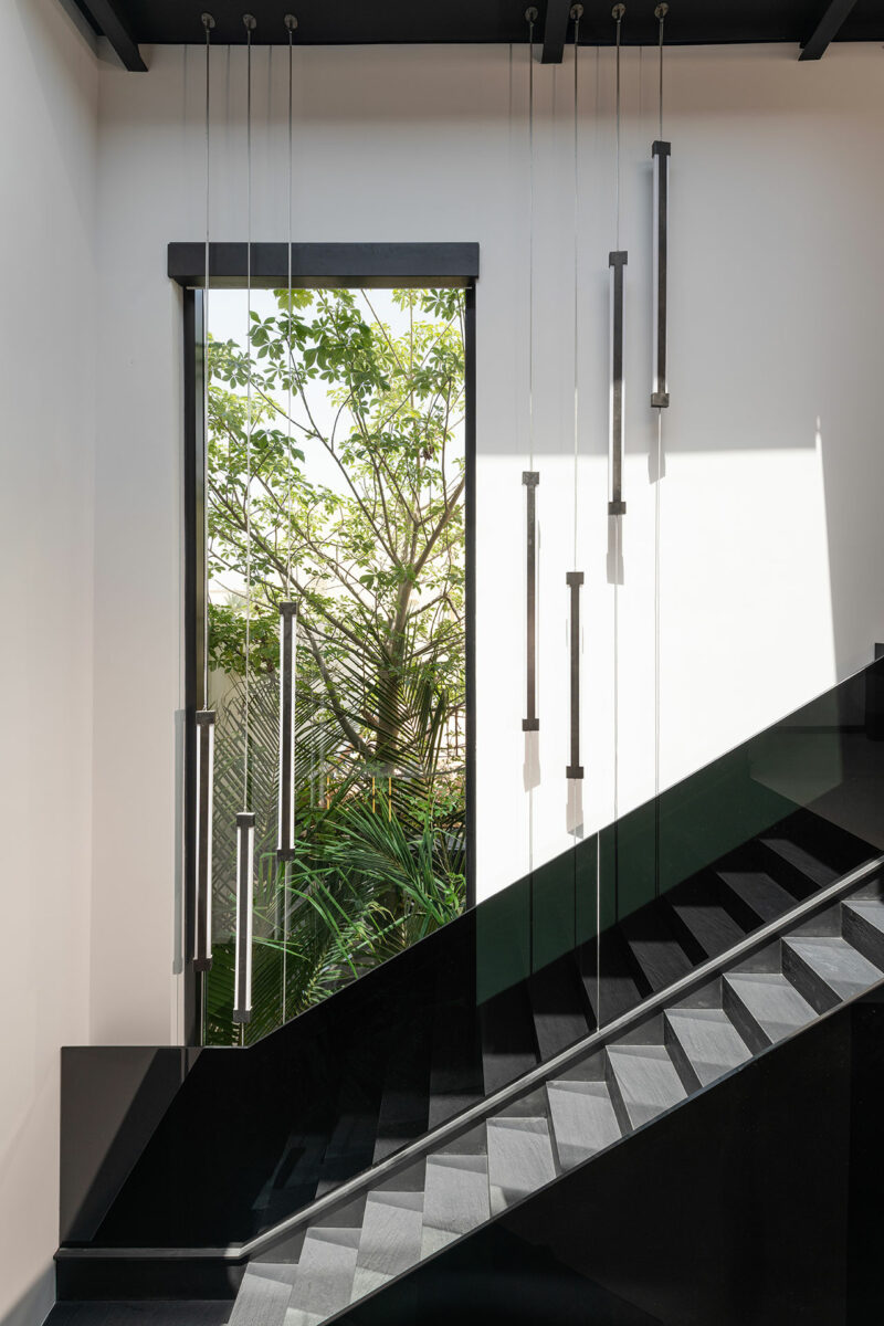 Modern staircase with black steps and railing, vertical pendant lights, and a large window revealing green trees outside. Natural light enters through the window.