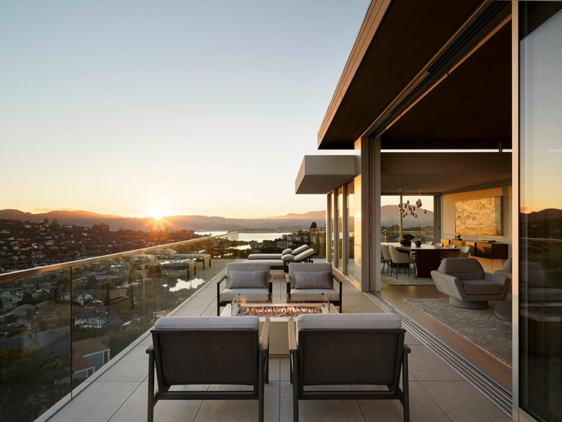 Modern outdoor terrace with seating area, glass railing, and a firepit, overlooking a city and water at sunset. Interior living room is visible through large open sliding doors.