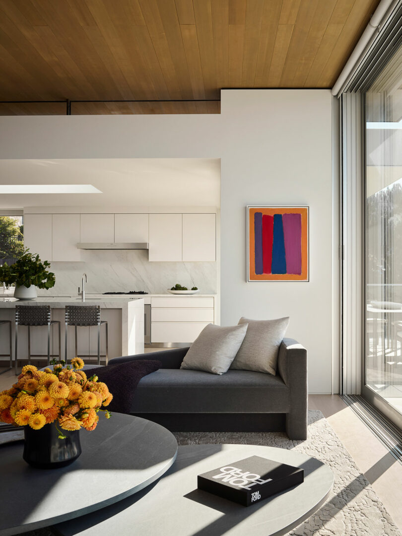Modern living room with a gray chaise lounge, round coffee table, vase of orange flowers, abstract wall art, and a view into a white kitchen with marble backsplash and bar stools.