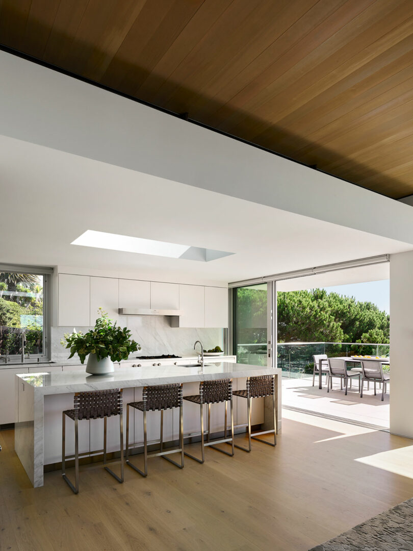 Modern kitchen with white cabinetry, marble island, four woven barstools, large windows, and sliding doors opening to an outdoor dining area with trees visible outside.