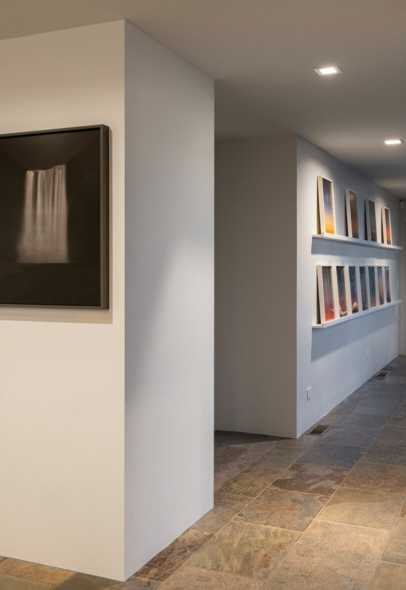 A hallway with framed photographs displayed on white walls, featuring tile flooring and recessed ceiling lights.