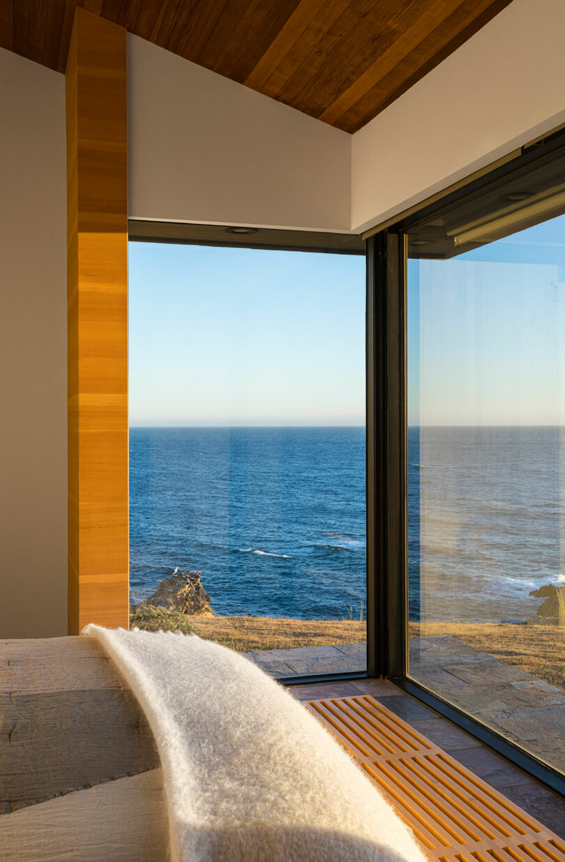 Modern bedroom with large floor-to-ceiling windows offering an unobstructed view of the ocean and rocky coastline at sunset.