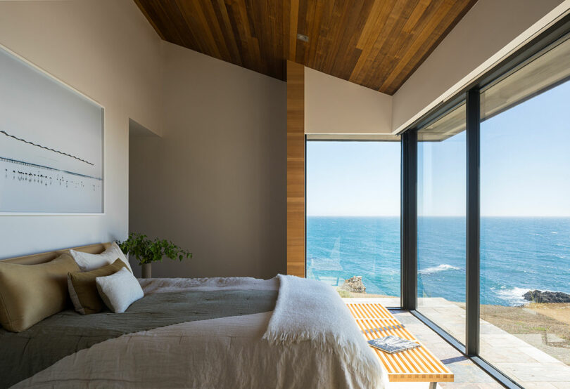Modern bedroom with large floor-to-ceiling windows offering an ocean view, a bed with neutral bedding, wooden ceiling, and minimalist decor.