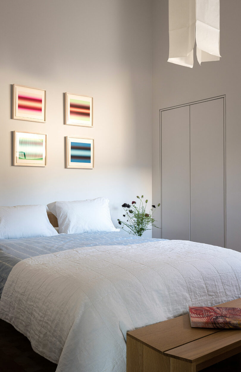 A neatly made bed with white bedding, a small vase of flowers on a side table, four colorful abstract artworks on the wall, and a closed closet door in a minimalist bedroom.
