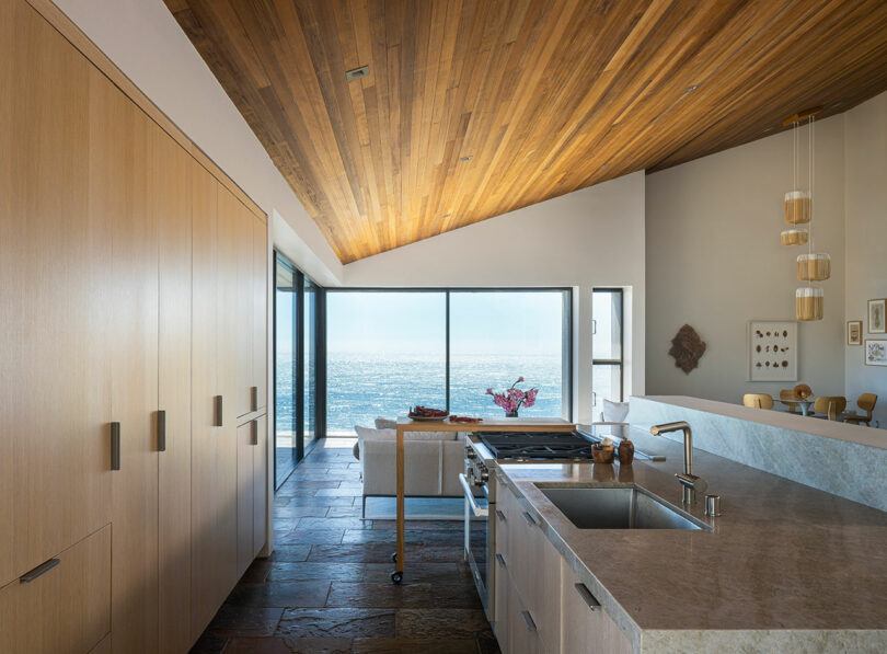 Modern kitchen with stone floors, wood-paneled ceiling, and large windows offering an ocean view. A small seating area and dining table are visible near the windows.