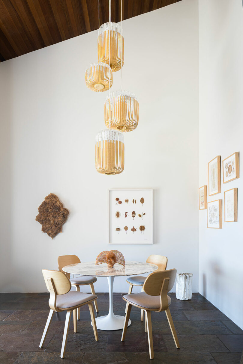 A round marble table with four chairs sits under a cluster of woven pendant lights; framed art and decor hang on white walls.