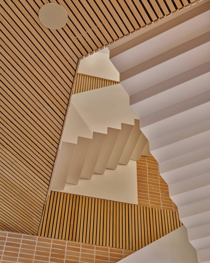 Geometric architectural interior featuring white staircases and ceilings with wooden slats and brick-patterned walls, viewed from below.