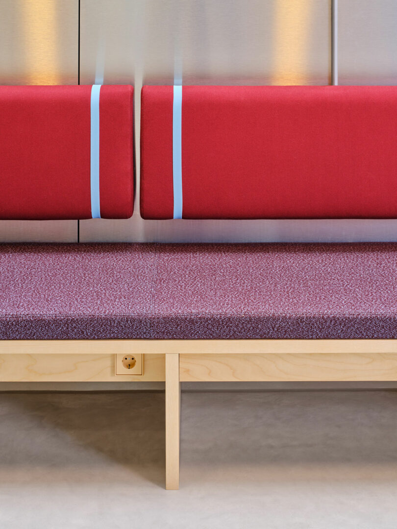 A bench with a wooden frame, purple speckled seat cushion, and two red back cushions with light blue straps against a metallic wall.