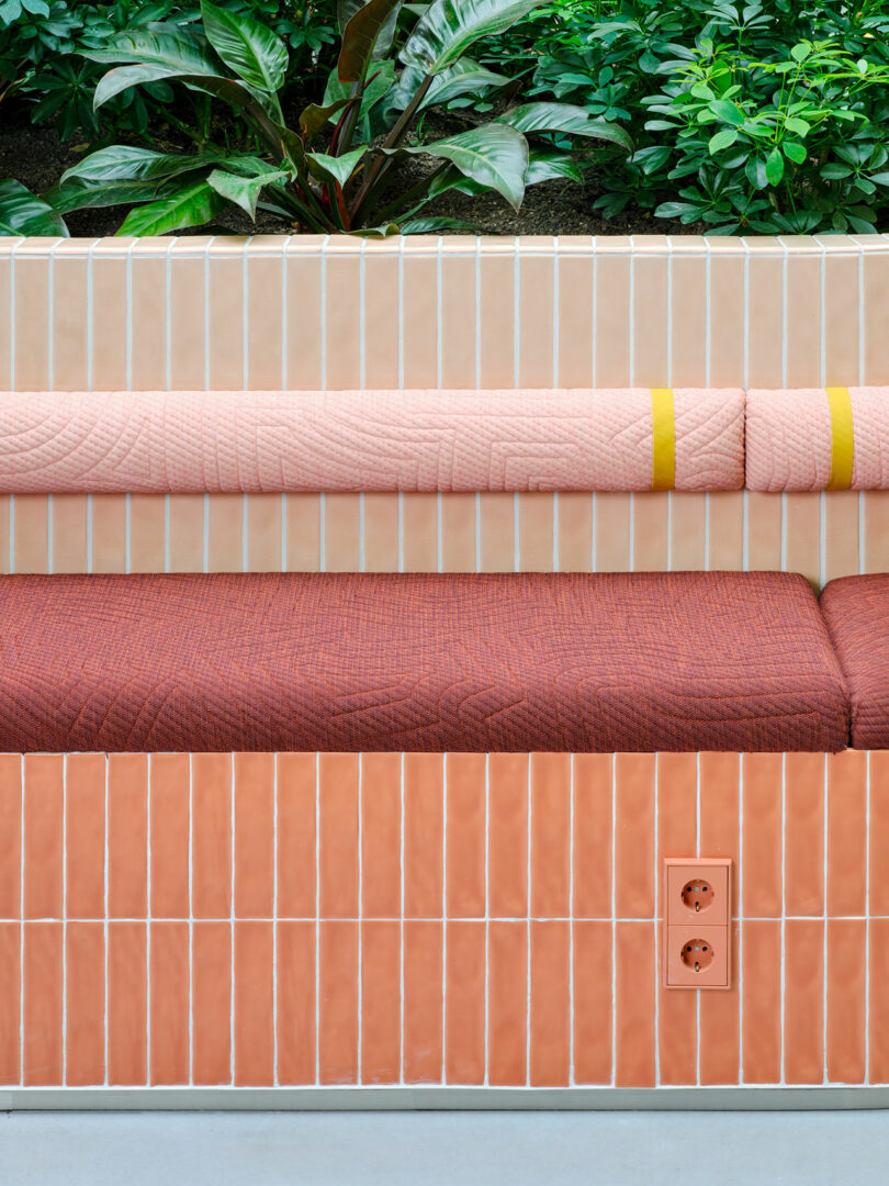 A tiled bench with pink and red cushions sits in front of a planter filled with green plants; a double power outlet is visible on the bench front.