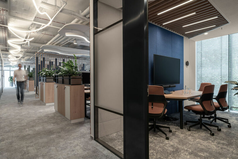 Modern open-plan office with BIT CREATIVE workstations, indoor plants, and a meeting area featuring a large screen and brown chairs. A person walks down the corridor on the left.