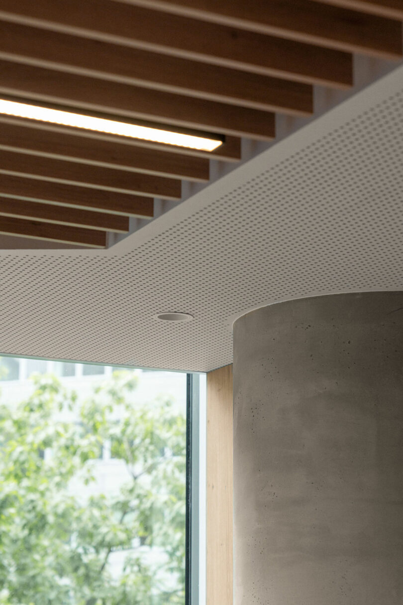 Modern BIT CREATIVE interior with a perforated white ceiling, exposed concrete column, wooden ceiling beams, and a large window that frames lush green trees outside.
