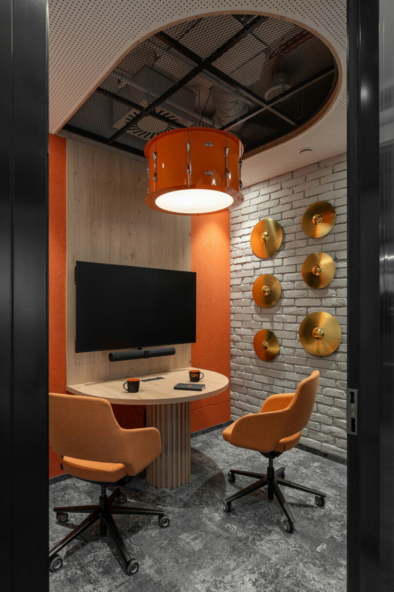 Small office meeting room by BIT CREATIVE, featuring two orange chairs, a round table, wall-mounted TV, drum-inspired ceiling light, and cymbals mounted on a white brick wall for decoration.