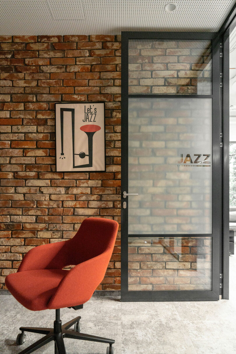 A modern office space by BIT CREATIVE features a red swivel chair, a brick wall, a framed "Let's Jazz" poster, and a glass door labeled "JAZZ.