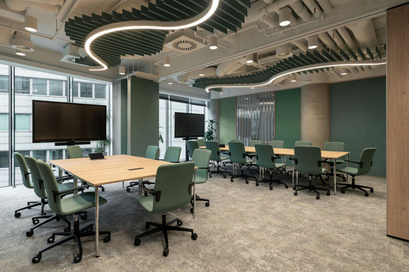 Modern conference room by BIT CREATIVE with several rectangular tables, green chairs on wheels, large windows, two flat-screen monitors, and wavy ceiling panels with exposed ducts.