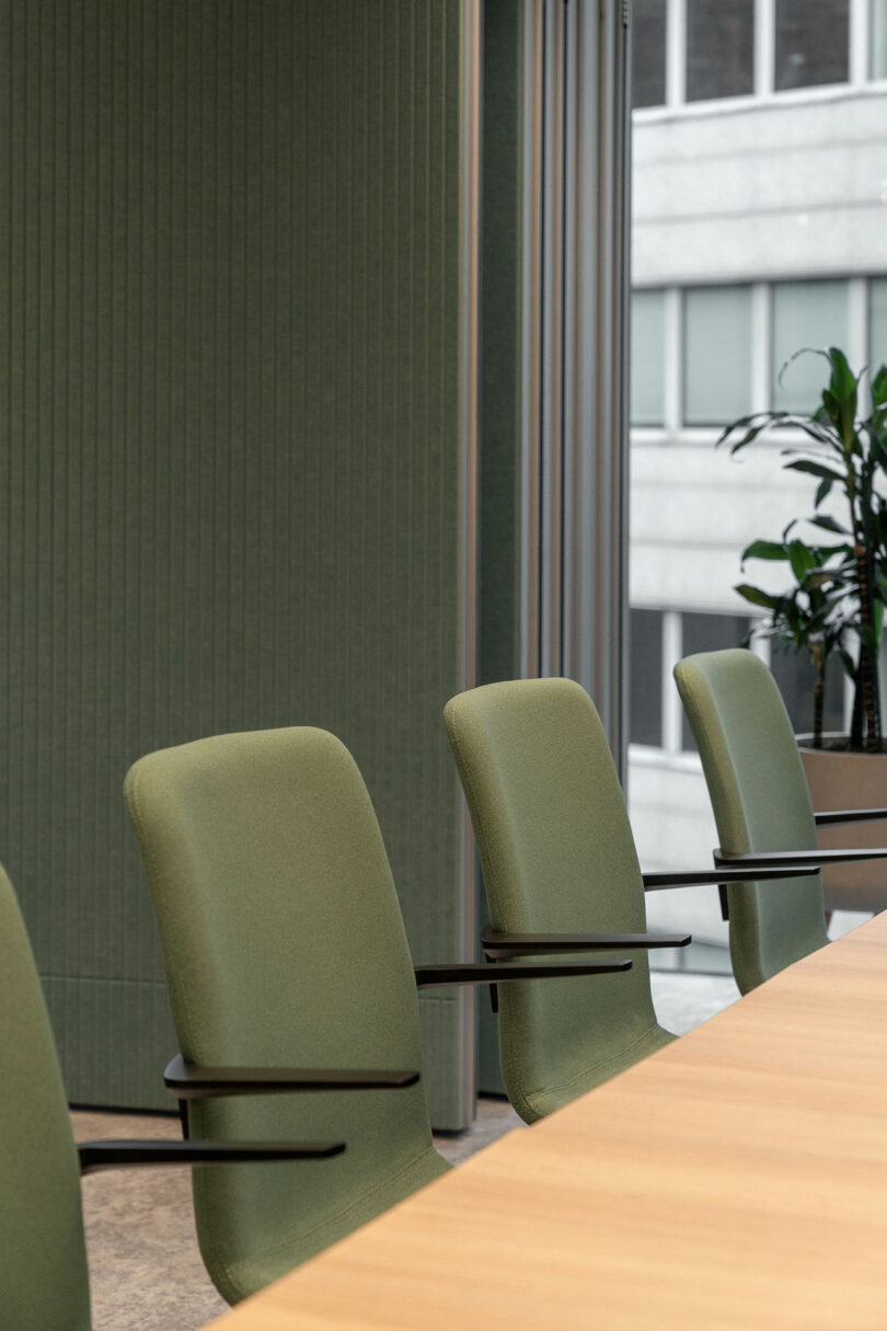 Three green office chairs are arranged around a light wood conference table in a modern BIT CREATIVE meeting room with large windows and a potted plant.