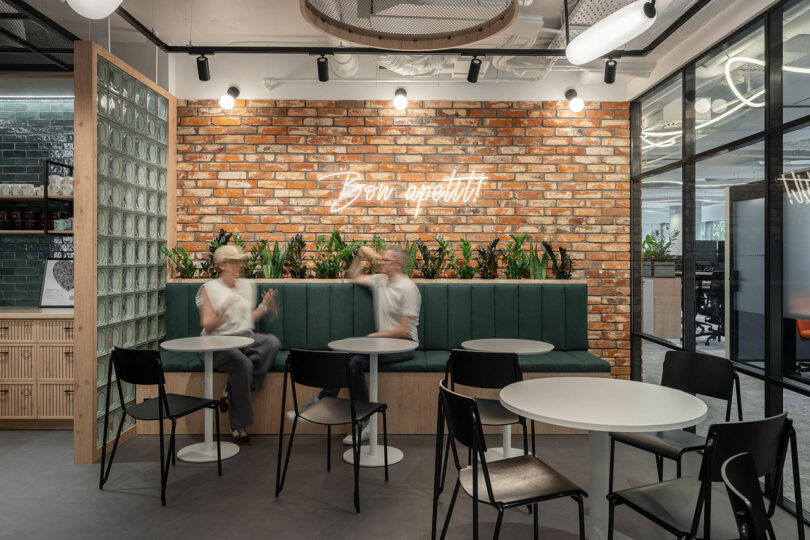 Modern office break area designed by BIT CREATIVE, featuring round tables, black chairs, green bench seating, a brick wall, neon "Bon appetit!" sign, and two people conversing.