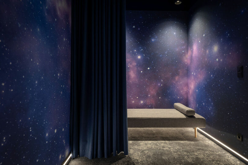 A BIT CREATIVE small room with walls and ceiling decorated in a galaxy pattern, featuring a single gray bench, a cylindrical pillow, and a dark curtain partially covering one side.