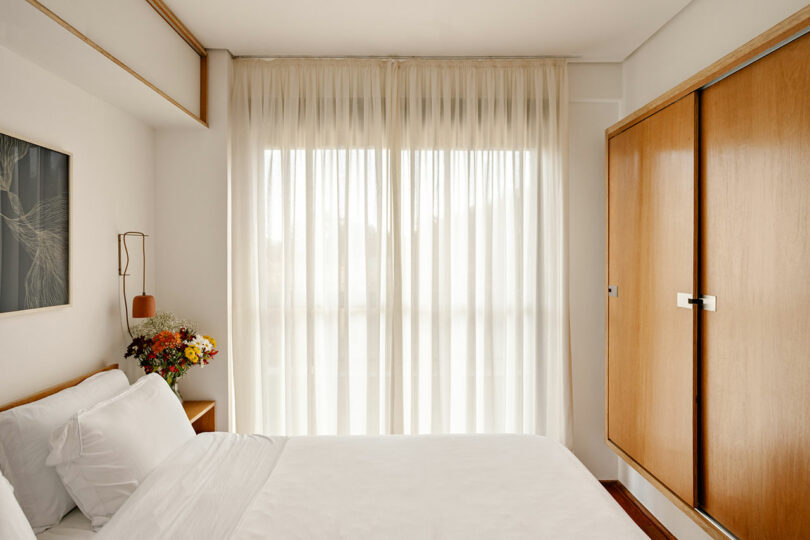A minimalist bedroom with a neatly made bed, white linens, wooden cabinets, a bouquet of flowers on the nightstand, and sheer curtains letting in natural light.