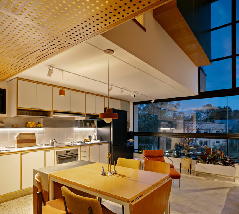 Modern kitchen and dining area with wood and neutral tones, large windows showing a nighttime view, and minimalist furniture.