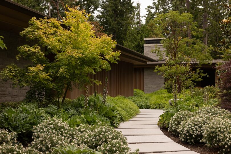 A modern house with stone and wood exterior is surrounded by lush green landscaping, featuring a curving concrete walkway and various trees and shrubs.