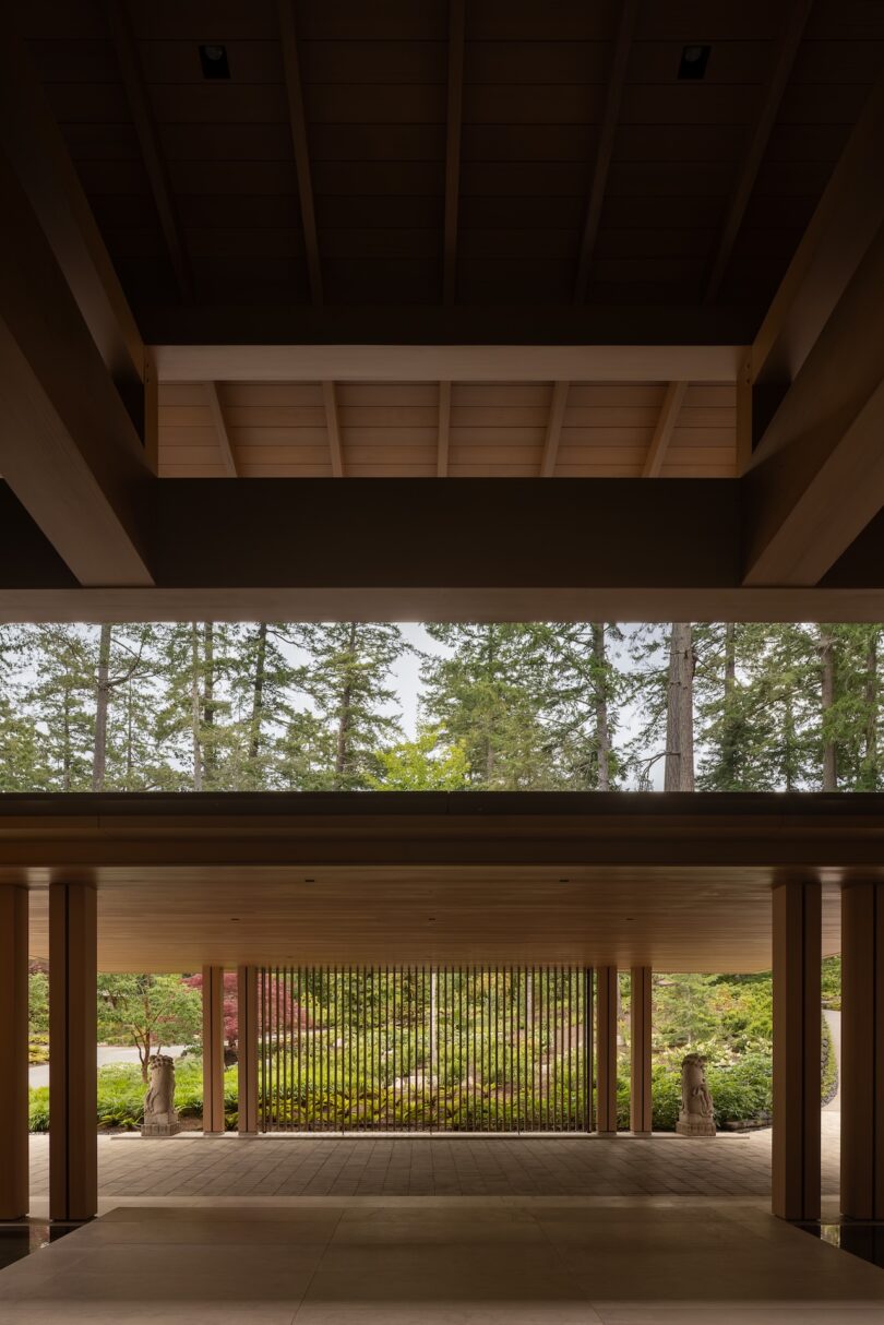 A view through a modern wooden structure with open beams and columns, looking out to a garden and tall trees beyond.