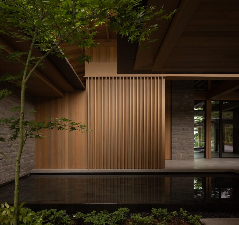 A modern architectural space with vertical wooden slats, stone walls, a shallow reflecting pool, and green plants in the foreground.