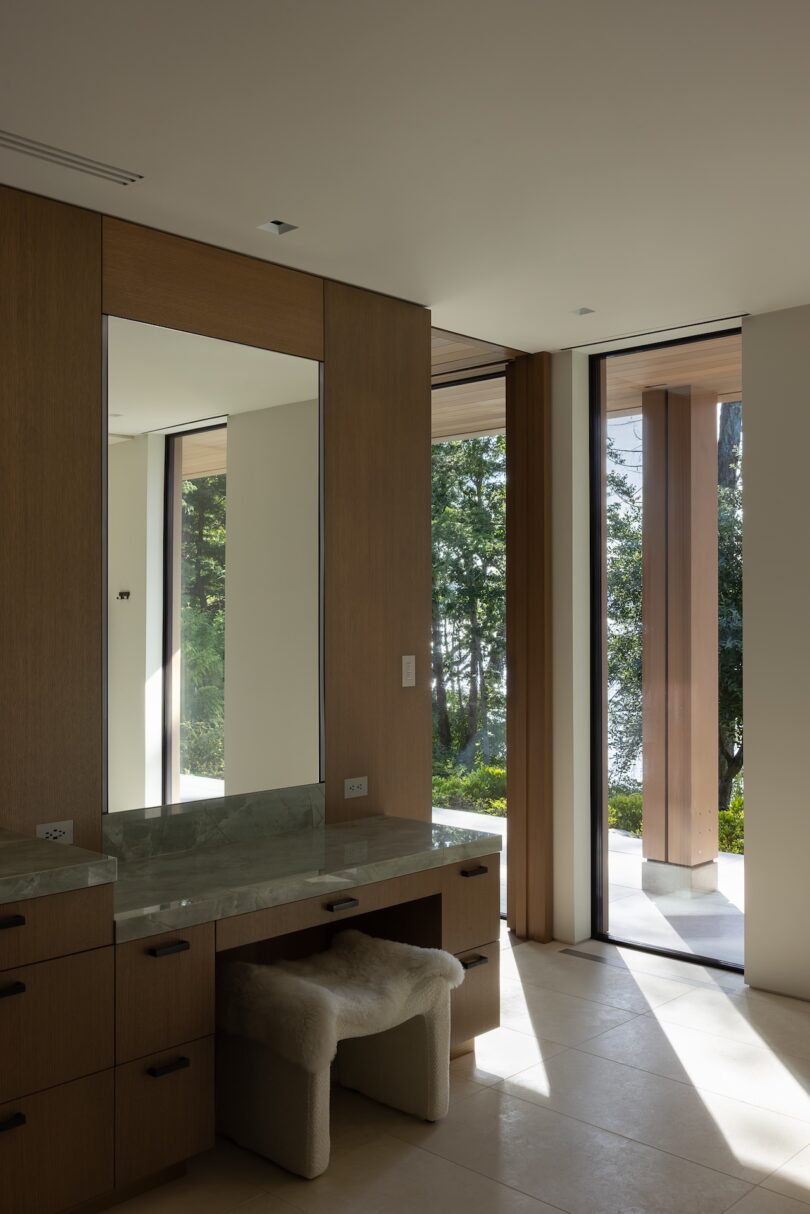 A modern bathroom with a marble-topped vanity, a plush stool, wood cabinetry, large mirror, and floor-to-ceiling windows overlooking greenery outside.